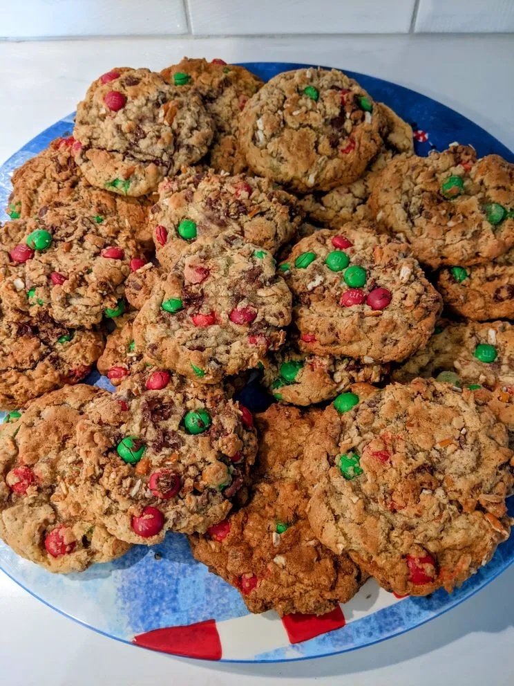 A winter blue plate with 22 perfectly baked, golden brown, holiday monster cookies stacked two layers deep, each busting with chocolate chips and chunks, red and green M&Ms, and broken apart pretzels.