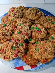 A winter blue plate with 22 perfectly baked, golden brown, holiday monster cookies stacked two layers deep, each busting with chocolate chips and chunks, red and green M&Ms, and broken apart pretzels. (opens in a new tab)